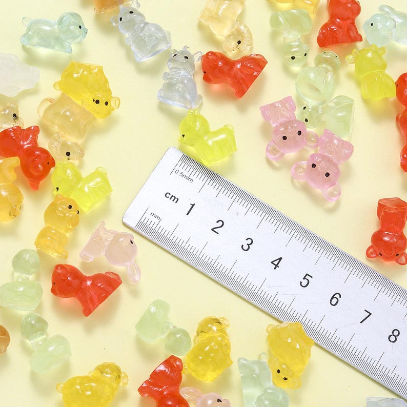 May include: A collection of small, colorful animal-shaped resin charms in shades of red, yellow, pink, and clear. A ruler is included for size reference. The charms are scattered on a light yellow surface.