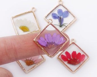 May include: Four gold-toned square pendants with different pressed flowers inside. The pendants are on a white surface.