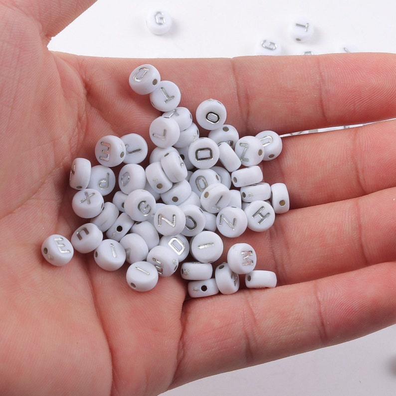 May include: A collection of white, round beads with silver letters and numbers. The beads are scattered in a person's hand, with some loose beads in the background. The beads are for jewelry making or crafting.