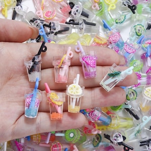 May include: A hand holding several miniature plastic drink cups with straws. The cups are clear with colorful contents and fruit slices. Some cups have pink, blue, and white liquids. The straws are black, white, and blue. The drinks are a fun, colorful assortment.