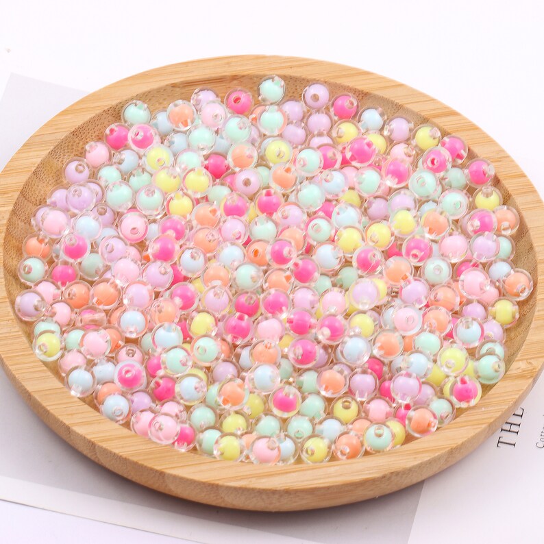May include: A wooden bowl filled with a multitude of colorful, translucent beads. The beads are in shades of pink, orange, yellow, green, and blue. These small, round beads are ideal for jewelry making and craft projects.