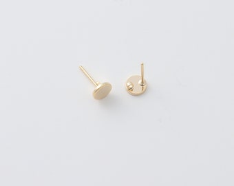 May include: Two gold-colored stud earrings with a simple, minimalist design. Each earring features a small, flat, circular disc and a post for insertion into the ear. The earrings are set against a plain white background.