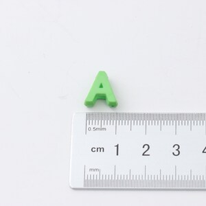 May include: A green plastic letter A, measuring approximately 1 cm in height, is placed next to a ruler.