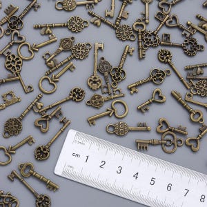 May include: A collection of antique bronze-colored keys with various decorative designs, including heart shapes and ornate patterns. A ruler with centimeter markings is in the image. The keys are scattered across a gray surface.
