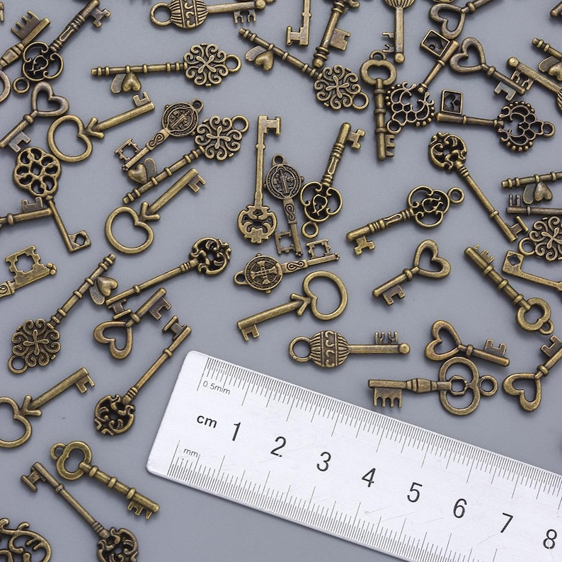 May include: A collection of antique bronze-colored keys with various decorative designs, including heart shapes and ornate patterns. A ruler with centimeter markings is in the image. The keys are scattered across a gray surface.