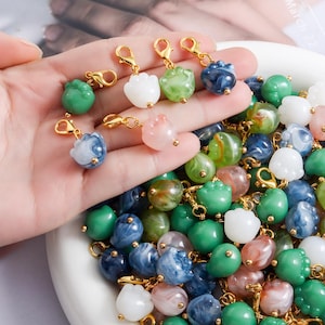 Puede incluir: Una colección de coloridos abalorios con cierres dorados. Los abalorios son de varios colores, incluyendo verde, blanco, azul y rosa, y tienen forma de pequeñas flores o capullos. Los abalorios se muestran en un cuenco blanco y se sostienen en una mano.