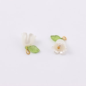 Puede incluir: Dos abalorios de flores blancas con hojas verdes y detalles dorados. Los abalorios tienen forma de campana y son perfectos para a&ntilde;adir un toque de elegancia a las joyas o las manualidades.