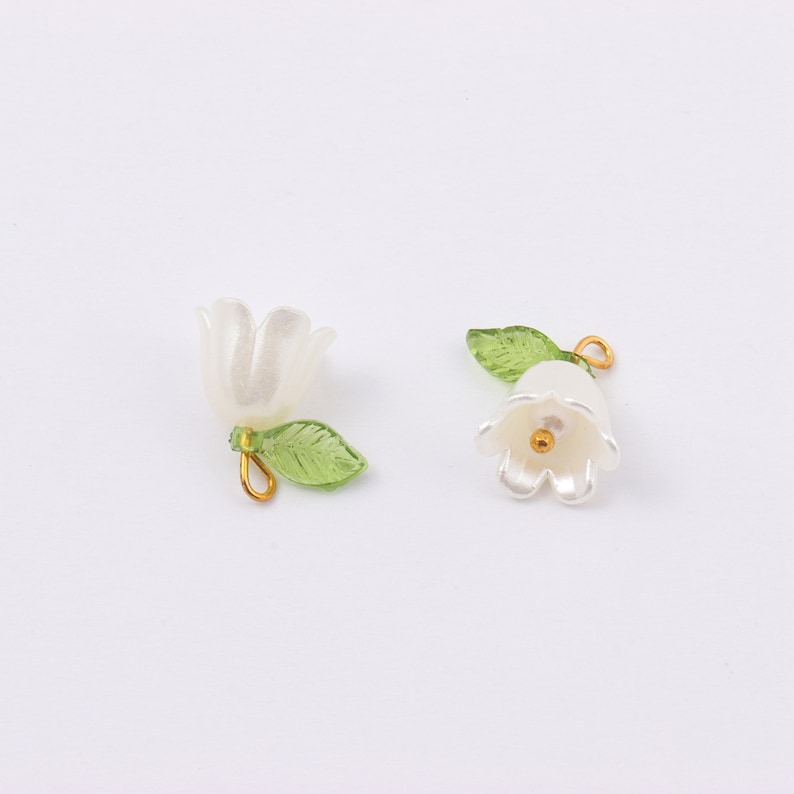 Puede incluir: Dos abalorios de flores blancas con hojas verdes y detalles dorados. Los abalorios tienen forma de campana y son perfectos para a&ntilde;adir un toque de elegancia a las joyas o las manualidades.