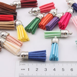 May include: Assortment of colourful suede tassels with silver metal caps. The tassels are approximately 5 cm long and are arranged on a white surface. A ruler is in the bottom of the image with measurements in centimetres.