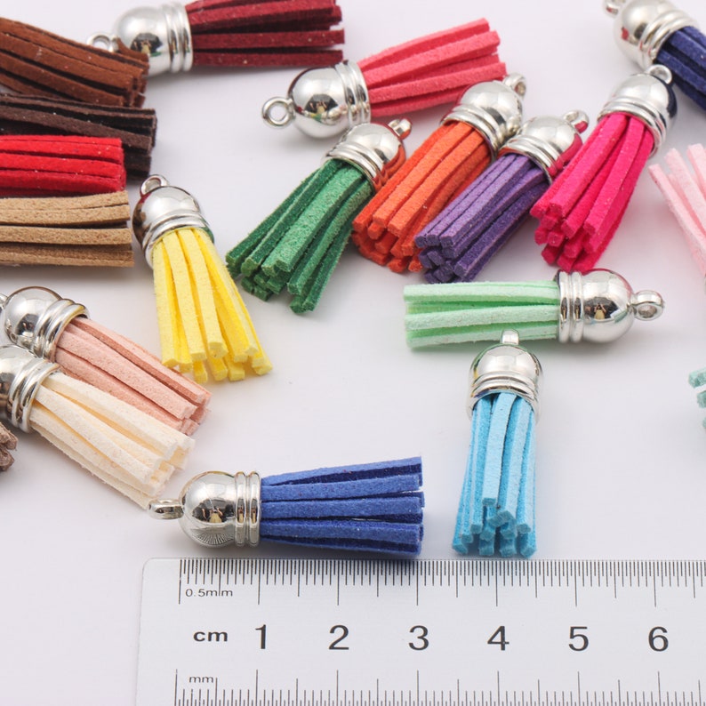 May include: Assortment of colourful suede tassels with silver metal caps. The tassels are approximately 5 cm long and are arranged on a white surface. A ruler is in the bottom of the image with measurements in centimetres.