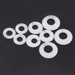 May include: A collection of white, circular pendants with a hole in the center. The pendants vary in size, with a pearlescent sheen. Each pendant has a small hole at the top for stringing. The background is a solid black.