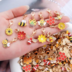 May include: A collection of gold-tone metal charms featuring enamel designs of bees, sunflowers, and honeycombs. Each charm is equipped with a small lobster clasp. The designs include yellow and black bees, red and yellow flowers, and orange honeycombs.