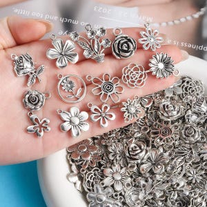 May include: A collection of silver-colored flower charms in various designs, including roses, daisies, and abstract floral shapes. Some charms have loops for attaching to jewelry or crafts. The charms are displayed in a white bowl.