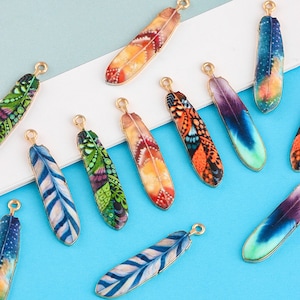 以下が含まれることがあります： 金色の金属ループが付いたカラフルな羽根チャームの詰め合わせ。各チャームは、青、緑、オレンジ、紫などの鮮やかな色で、異なるパターンデザインが施されています。