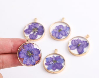 May include: Five round pendants with gold-colored frames. Each pendant features a pressed purple flower encased in clear resin. The pendants are displayed on a white background, showcasing their delicate floral designs.