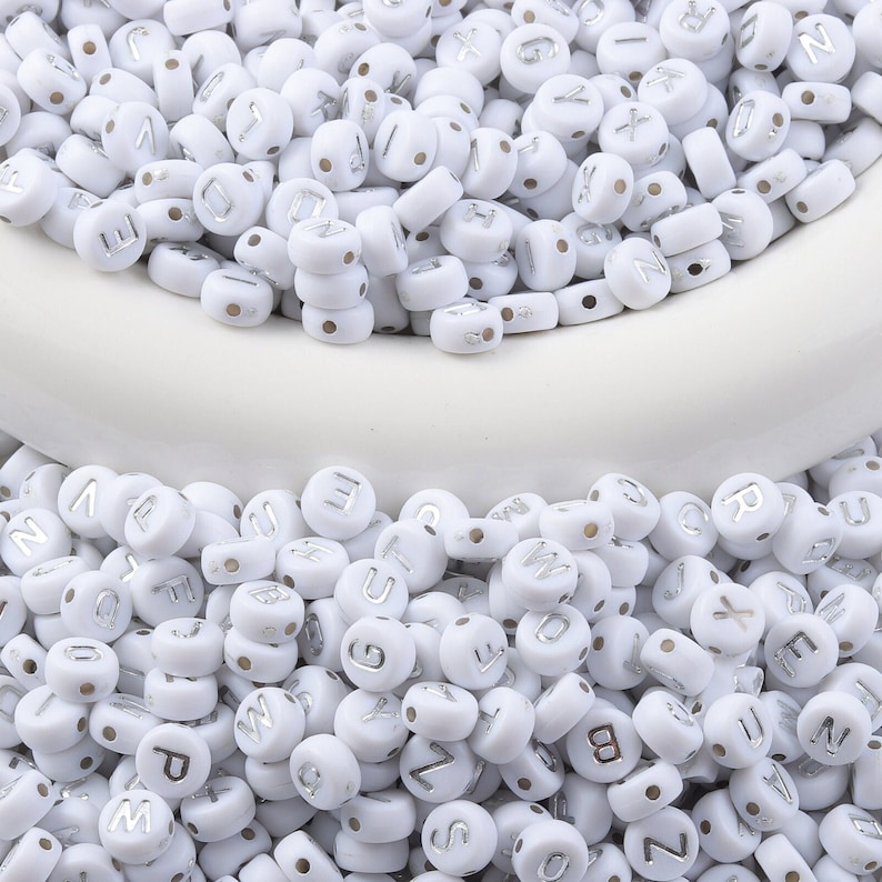 May include: White round beads with silver letter imprints, scattered across a white surface. The beads are uniform in size and shape, with a small hole for stringing. Ideal for jewelry making and crafts.