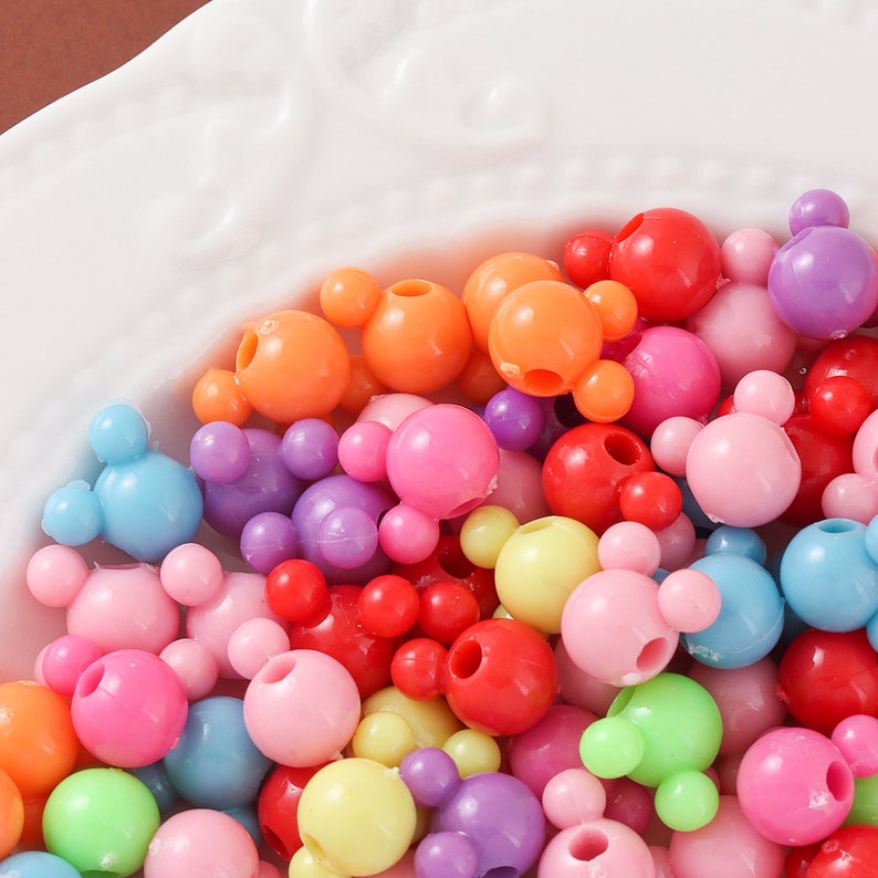 May include: A collection of colorful plastic beads shaped like Mickey Mouse heads. The beads are in various shades of pink, purple, blue, green, yellow, orange, and red.