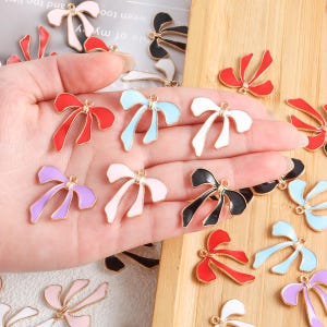 Peut inclure: Une collection de breloques en forme de nœuds colorés, dans diverses couleurs, dont le rouge, le noir, le blanc, le rose et le bleu. Chaque breloque a un contour doré et une petite boucle pour l'attacher à des bijoux ou autres créations. Les breloques sont présentées sur une surface en bois.