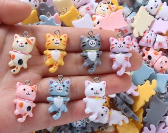 Puede incluir: Una colección de colgantes con forma de gato de colores, en tonos naranja, rosa, azul y blanco. Cada colgante tiene un pequeño lazo metálico para sujetarlo a joyas o manualidades. Los colgantes están en varias poses y tienen expresiones faciales lindas.