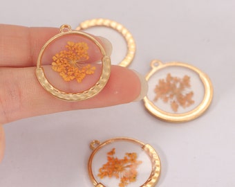 May include: Gold-colored round pendants with a clear resin center, each containing a dried orange flower. The pendants have a hammered texture on the bottom half of the frame. The background is a light, neutral color.