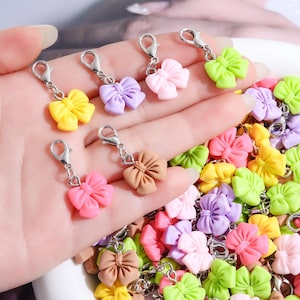 Pode incluir: Uma coleção de pequenos pingentes em forma de laço em várias cores pastel, incluindo amarelo, lavanda, rosa claro, verde-limão e bege. Cada laço está preso a um fecho lagosta de metal prateado, projetado como acessório decorativo.