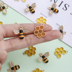 Puede incluir: Amuletos dorados de abeja y panal con detalles de esmalte negro y amarillo. Los amuletos de abeja tienen alas de pedrería. Los amuletos están sobre un fondo verde claro y blanco.