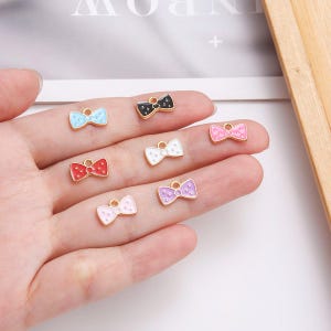 Peut inclure: Plusieurs breloques en forme de nœud papillon de différentes couleurs : bleu clair, noir, rouge, blanc, rose et violet. Chaque breloque a un bord doré et une petite boucle pour l'attache. Les nœuds sont ornés de petits pois blancs.