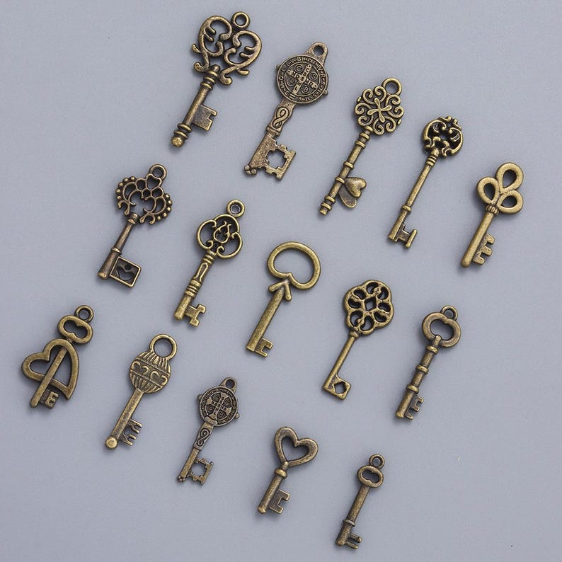 May include: An assortment of antique-style keys in various designs. The keys are made of a bronze-colored metal and feature ornate details, including floral patterns, hearts, and religious symbols. The keys are arranged on a light gray surface.