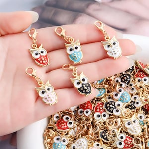 Pode incluir: Uma coleção de pingentes de coruja em metal dourado com detalhes em esmalte nas cores vermelho, azul, branco, rosa e preto. Cada pingente de coruja tem olhos brancos grandes com pupilas pretas e um pequeno fecho lagosta no topo.
