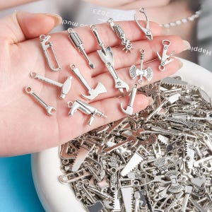 May include: A collection of silver-colored miniature tool charms, including hammers, saws, wrenches, and screwdrivers. The charms are held in a hand and also in a white bowl. These charms are ideal for jewelry making or craft projects.