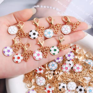 May include: A collection of gold-tone metal charms featuring a soccer ball design and the year 2026. Each charm includes a lobster clasp and a soccer ball with various colored panels, including red, blue, green, purple, pink, and black and white.