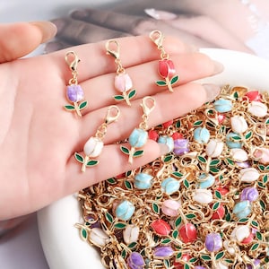 May include: A collection of gold-tone metal tulip charms with enamel petals in shades of red, pink, white, purple, and light blue. Each charm features a green stem and leaf detail, attached to a small gold lobster clasp for easy attachment.