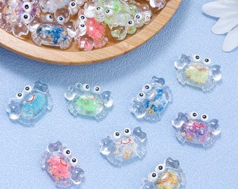 May include: Clear resin crab charms with colourful fillings, such as blue, green, pink, and purple. The charms have smiling faces and are scattered on a light blue surface and in a wooden bowl.