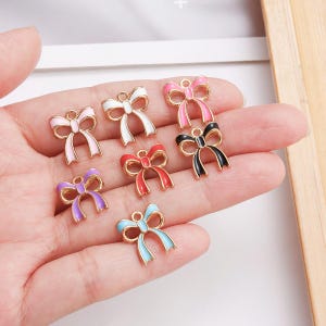 May include: A collection of colorful bow-shaped charms in various colors, including pink, white, red, purple, black, and blue. Each charm has a gold-colored outline and a loop for attaching to jewelry or other crafts.