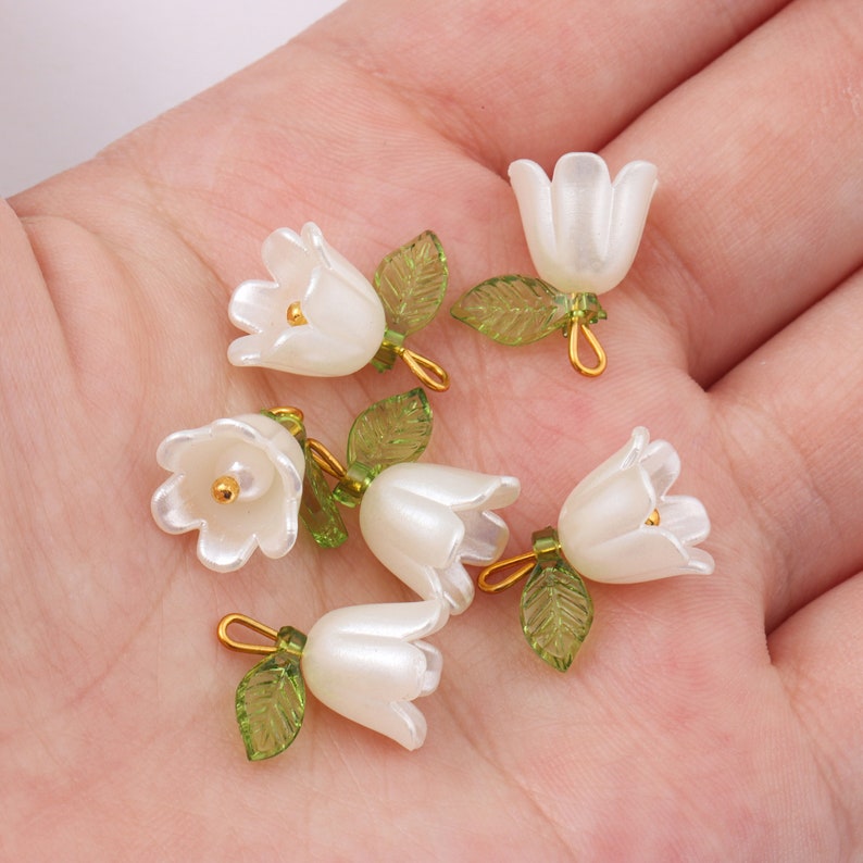 Puede incluir: Una colecci&oacute;n de seis abalorios de flores blancas con hojas verdes y detalles dorados. Los abalorios est&aacute;n hechos de pl&aacute;stico y tienen un acabado perlado.