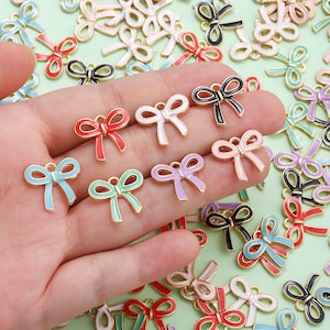 May include: A selection of enamel bow charms in a range of colours, such as red, blue, green, and pink. Each charm has a gold-coloured outline and a small loop for attaching to jewellery or crafts. The bows are in a variety of colours.