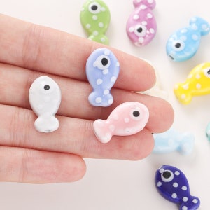 Pode incluir: Conjunto de contas de peixe de cer&acirc;mica coloridas com bolinhas brancas e olhos pretos. As contas s&atilde;o de v&aacute;rias cores, incluindo azul, rosa, verde, amarelo e branco.