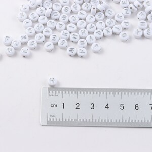 May include: White round beads with silver letter imprints scattered across a white surface. A ruler with centimeter and millimeter markings is in the foreground. The beads are approximately 0.5 cm in diameter, perfect for jewelry making or crafts.
