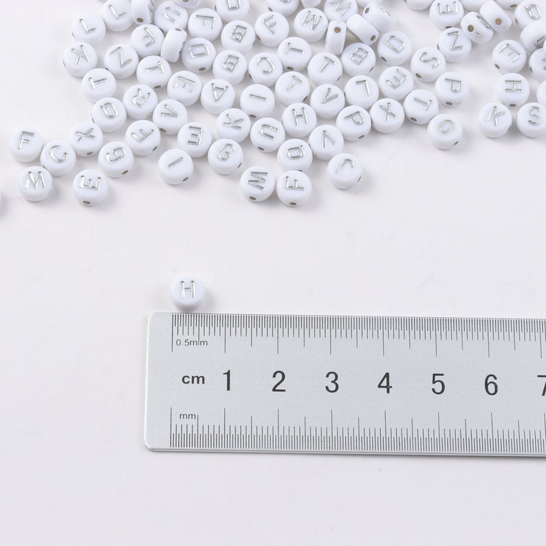 May include: White round beads with silver letter imprints scattered across a white surface. A ruler with centimeter and millimeter markings is in the foreground. The beads are approximately 0.5 cm in diameter, perfect for jewelry making or crafts.
