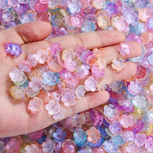 Pode incluir: Um conjunto de pequenas contas em forma de flor em cores pastel, incluindo rosa, roxo, azul e amarelo. Estas contas translúcidas são ideais para artesanato ou criação de joias.