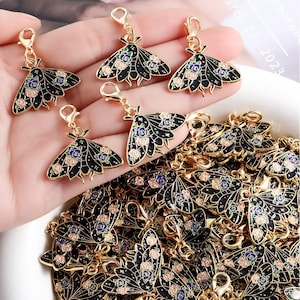 Puede incluir: Encantos de metal dorado con forma de polilla con alas de esmalte negro y detalles florales en rosa y azul. Los amuletos tienen un cierre de mosquetón para una fácil fijación. Colección de amuletos decorativos.