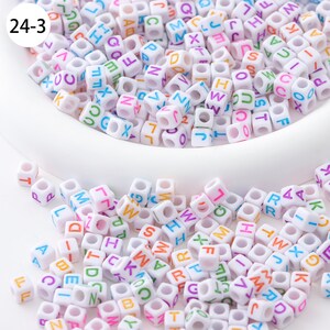 May include: A close-up of a white bowl filled with white plastic letter beads. The beads are in various colors and have a hole in the center. The beads are scattered around the bowl on a white surface.