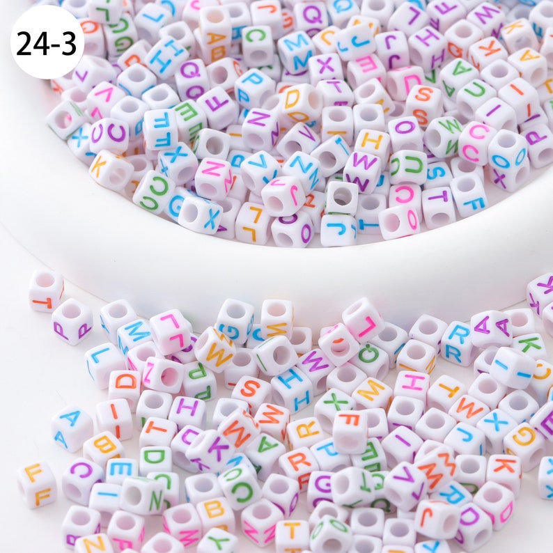 May include: A close-up of a white bowl filled with white plastic letter beads. The beads are in various colors and have a hole in the center. The beads are scattered around the bowl on a white surface.