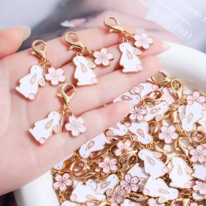 Puede incluir: Una colección de dijes de metal en tono dorado con un conejo de esmalte blanco y una flor de cerezo de esmalte rosa claro. Cada dije está unido a un broche de langosta dorado, creando un conjunto de accesorios decorativos para joyería.