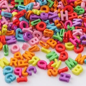 May include: A large pile of colorful plastic letter beads in various colors, including red, orange, yellow, green, blue, purple, and pink. The beads are arranged in a random pattern and are scattered across a white surface.