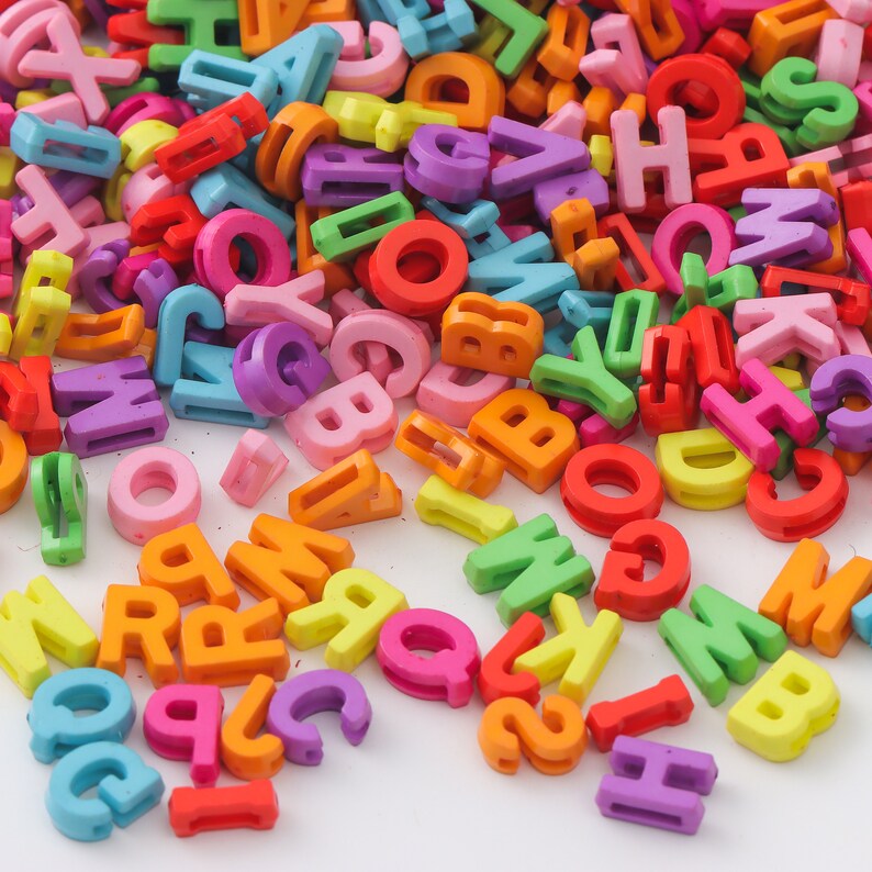 May include: A large pile of colorful plastic letter beads in various colors, including red, orange, yellow, green, blue, purple, and pink. The beads are arranged in a random pattern and are scattered across a white surface.
