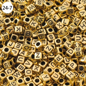 May include: A close-up image of a large pile of gold-colored letter beads. The beads are cube-shaped and each has a black letter printed on one side.
