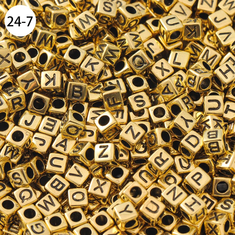 May include: A close-up image of a large pile of gold-colored letter beads. The beads are cube-shaped and each has a black letter printed on one side.