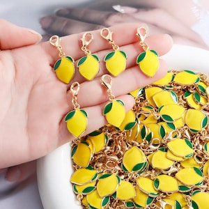Pode incluir: Pingentes de limão dourados com fruta de esmalte amarelo e detalhes de folhas verdes. Os pingentes têm um fecho de lagosta para fácil fixação. Uma coleção de pingentes semelhantes é visível no fundo.