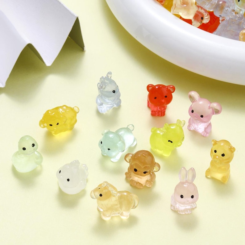 May include: A collection of small, translucent animal figurines in various colors, including yellow, red, pink, and green. The figurines depict a variety of animals, such as bears, rabbits, and sheep. The figurines are scattered on a light yellow surface.